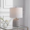 This table lamp has a concrete base with white etching, brushed nickel accents, and a light beige linen shade, offering modern sophistication and understated elegance.